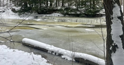 A frozen river flows through a snowy forest, featuring a small icy waterfall and snow-covered logs along the bank.