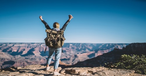 Back view of excited hipster girl with backpack raising hands on mountain top happy with achievement in hiking, rear view of female traveler happy feeling freedom high on rocky cliffs in grand Canyon