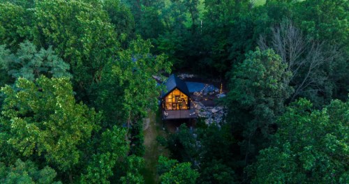 Aerial view of a cabin with glowing windows, surrounded by lush green trees and rocky terrain.