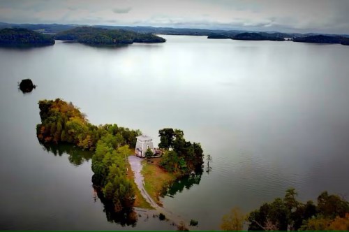 A lone island off the coast of the shoreline of this clear and calm lake with a lighthouse perched right in the center of the island.
