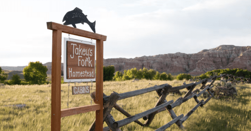 At the end of a rustic fence, a sign with the name of a ranch signals beside a rural road.