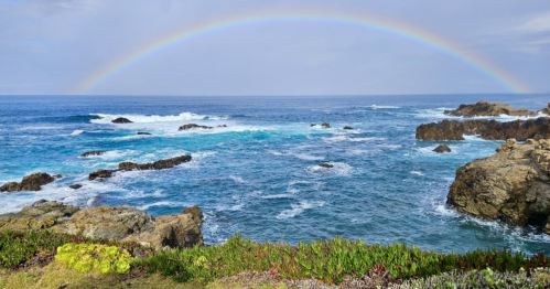 The Pacific Ocean crashes against a rugged, rock beach near Carmel-by-the-Sea while a rainbow stretches overhead.