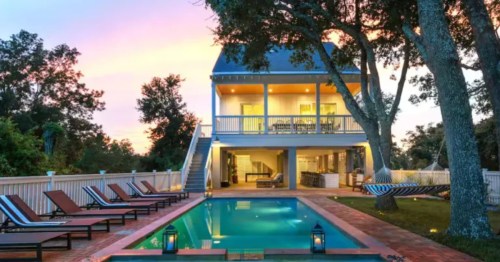 House with a pool and hot tub, lounge chairs, a hammock, and trees under a colorful sunset sky.