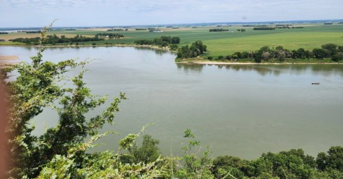 High-angle view of a wide river winding through green Nebraska farmland and lush trees under a clear, pale blue sky.