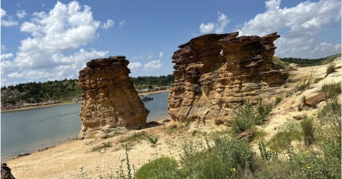 Dramatic golden rock pillars standing on a sandy beach beside a calm blue lake.
