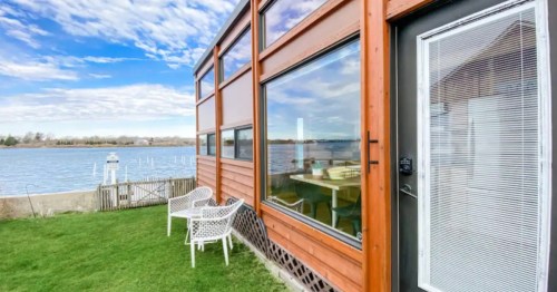 Modern wood-paneled tiny house with large windows, white patio chairs, and a stunning view of a calm blue river.