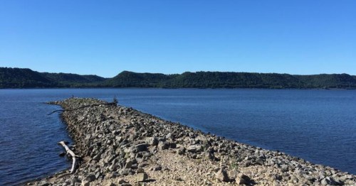 A long, rocky breakwater extends into a wide blue river, with lush green bluffs lining the horizon under a clear sky.