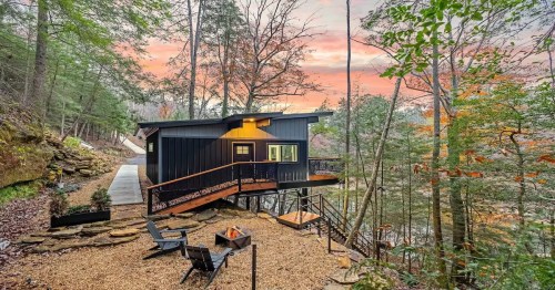 Elora's Cabin, a dark and wooden cabin in the woods of Alabama with two lawn chairs in the foreground and a pink-colored sky in the back