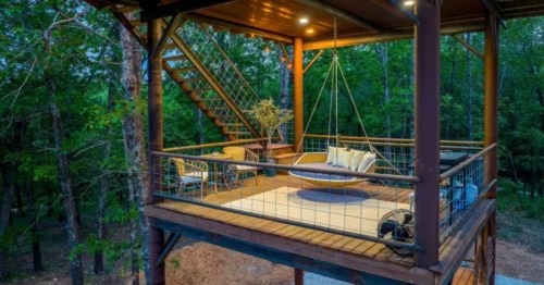 A cozy elevated wooden deck in a Missouri forest features a hanging daybed, wicker chairs, and string lights.