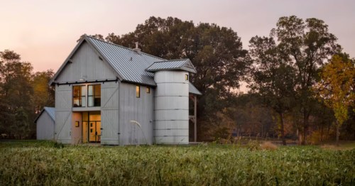 Modern grey barn-style house with a silo, large windows, and a metal roof surrounded by trees and a field.