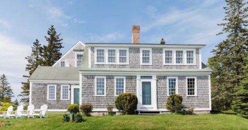 Gray shingle-sided house with white trim, a blue door, and white chairs on a green lawn under a blue sky.