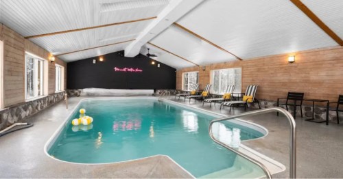 Indoor pool with lounge chairs. Sloped ceiling, wood walls, black accent wall with neon sign. Bright, airy space.
