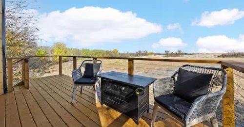 A wooden deck with two chairs and a fire table, overlooking open dunes and trees beneath a bright blue sky.