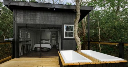 Modern treehouse painted black with a wooden deck, two white bathtubs, and a view into a bedroom with a bed.
