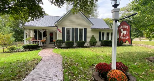 Views of the front of a charming Airbnb in Rocheport Missouri featuring a small porch with decorative flowers, a spacious yard, and surrounded by lush green trees.