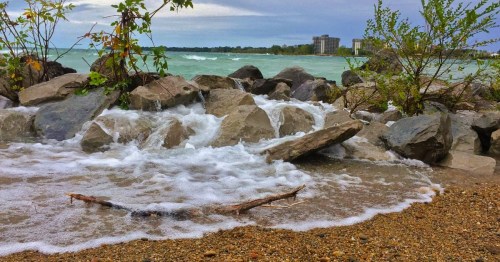 Waves crashing over rocks and sandy shore with a cloudy sky, at Port Huron, a spring destination in Michigan.