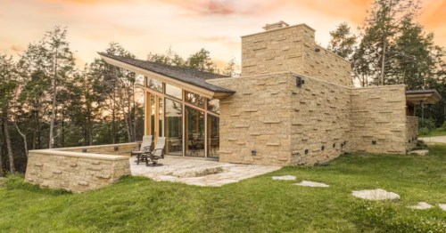 A stunning cottage, featuring stone masonry walls, a steeply angled roof, large windows, and a patio, with a sunset backdrop.