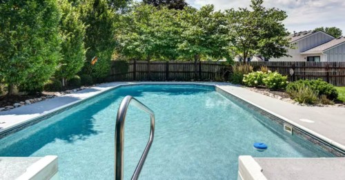 A rectangular swimming pool with clear blue water, surrounded by trees and a wooden fence.