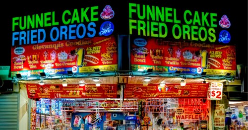 A colorful kiosk offering dozens of fair foods like funnel cakes and fried oreos, illuminated at night.
