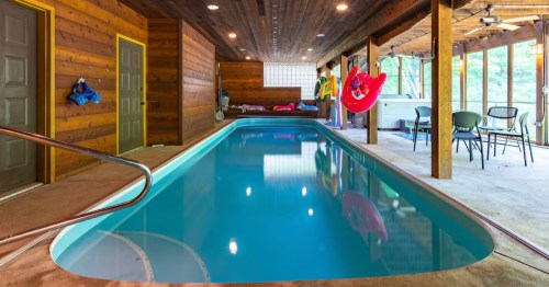 An indoor lap pool stretches under a wooden ceiling. Doors line one side, and a patio with chairs is visible beyond.
