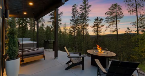 Outdoor patio with a fire table, chairs, a hanging swing, and a view of pine trees and a colorful sunset sky.