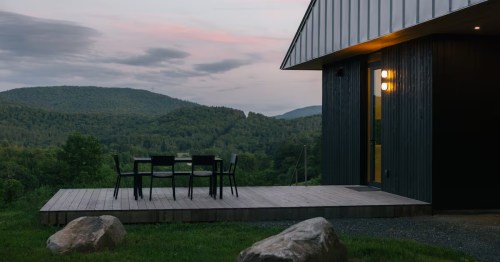 Gemini said Modern black cabin with a wooden deck overlooks lush green mountains under a soft, dusky pink and grey twilight sky.