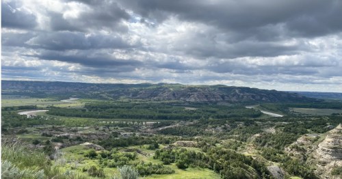 A winding river snakes through a lush green valley and badlands under a dramatic, cloudy sky.