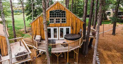 A wooden cabin on stilts among trees, with wraparound decks, large windows, and a hot tub nestled in the forest.