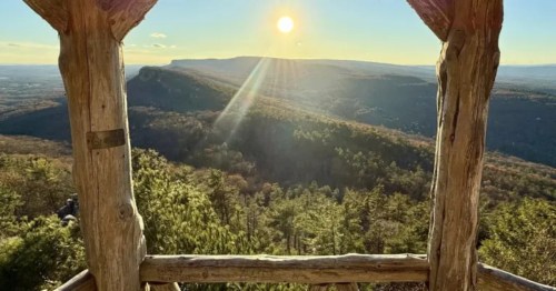A sunset view from outside a wooden gazebo, overlooking mountains and forests.