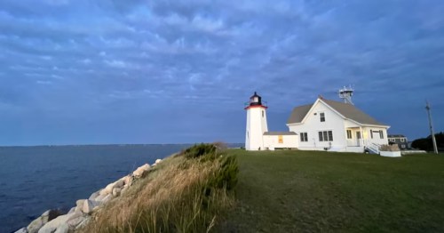 A white lighthouse and adjacent cottage sit on a grassy coastal point under a cloudy blue twilight sky next to the ocean.