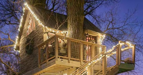 A cozy wooden treehouse with a wrap-around deck is illuminated by warm string lights against a purple twilight sky.