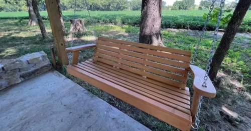 A wooden porch swing hangs invitingly under shady trees, overlooking a green field under a clear blue sky.