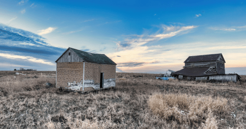 Two derelict, abandoned farm buildings in a dry, grassy field under a dramatic blue and orange sky.