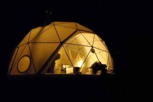 A glowing geodesic dome tent stands out against the dark night. Warm light spills from its windows.