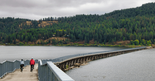 two people walking on a rail trail with a body of water and a forest of trees behind