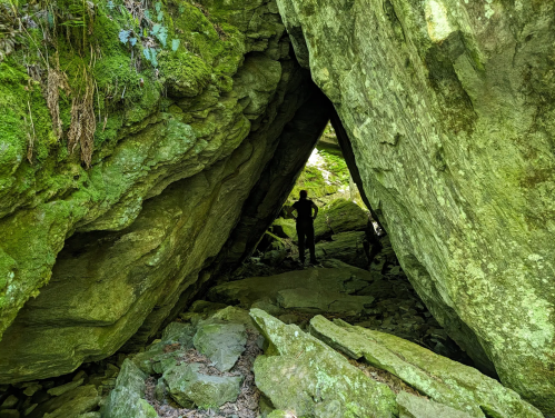 Moss-covered rocks form a triangular opening, framing a silhouetted figure in Devil's Gulch, Vermont.