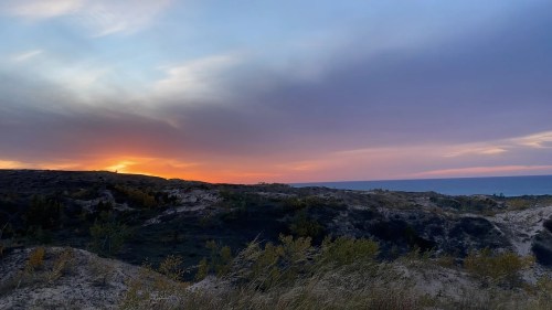 The sunset is visible behind rolling sand dunes and scrub bush, with Lake Michigan in the distance.