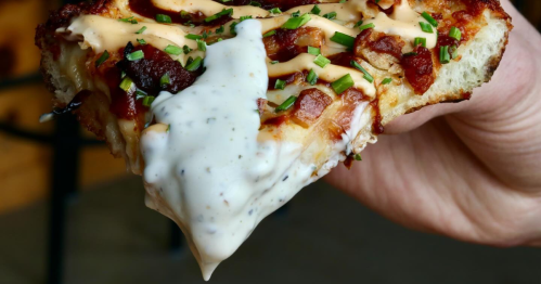 A diner holds up a slice of Sicilian-style pizza with pepperoni, dipped in a creamy ranch sauce.