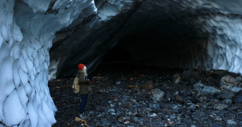A hiker in a red hat stands at the base of a towering ice cave.