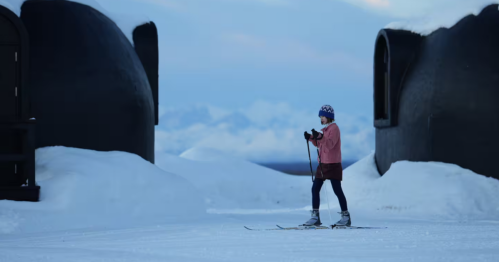 A woman in a pink coat cross-country skis between two buildings on a cold day.