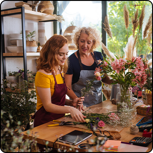 A mother and daughter stand at a work bench creating floral displays inside a plant store.