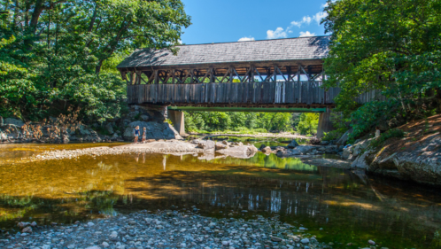 A side view of the Sunday River Covered Bridge in Newry, Maine on a sunny day. The bridge lies over the Sunday River. Two people can be seen on rocks in the water in front of the bridge