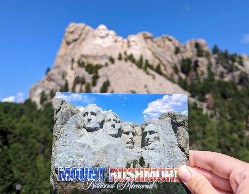 A hand holding a postcard depicting Mount Rushmore National Memorial in front of Mount Rushmore.