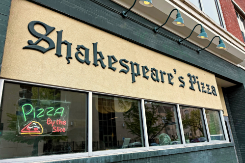 A storefront sign displays the name of Shakespeare's Pizza with a neon pizza sign in the window.