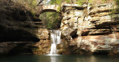 A pedestrian bridge on top of a waterfall that goes into a pond.
