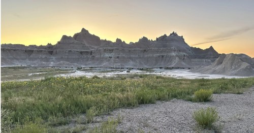 Jagged Badlands rock formations silhouetted against a golden sunset sky. A long boardwalk winds through the prairie.