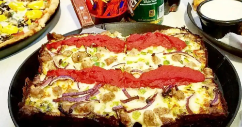 A rectangular Detroit-style pizza with cheese, green peppers, red onions, and red sauce. Drinks like Faygo and Vernors are in the background.