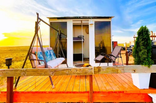 A tiny home Airbnb near Petrified Forest National Park, featuring large glass windows, a small wooden patio with a swinging bench, and views of the vast desert in the backdrop.