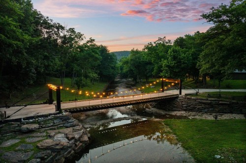 Views of a creek in Arkansas at sunset featuring a footbridge with string lights in a heavily wooded area