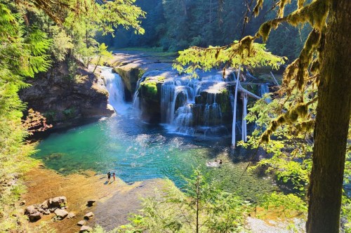 A wide waterfall flows into a turquoise pool, two people wade in the water and green trees surround the view.
